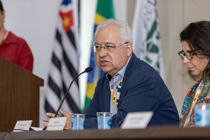 A imagem mostra Elbert Macau falando em um microfone durante um evento formal, sentado à mesa. Ele veste uma camisa azul claro com um casaco azul escuro e usa óculos. Há um crachá pendurado no pescoço, indicando sua participação oficial no evento. À sua frente, na mesa, estão copos de água e uma placa de identificação com seu nome, embora o texto não esteja completamente legível na imagem.  À sua direita, aparece parcialmente uma mulher, também sentada, com cabelo solto e óculos, olhando para baixo. Ao fundo, é possível ver bandeiras, incluindo a do Brasil e do estado de São Paulo, além de um púlpito à esquerda com parte de outra pessoa visível, vestindo vermelho. O ambiente sugere um painel ou conferência com caráter formal.