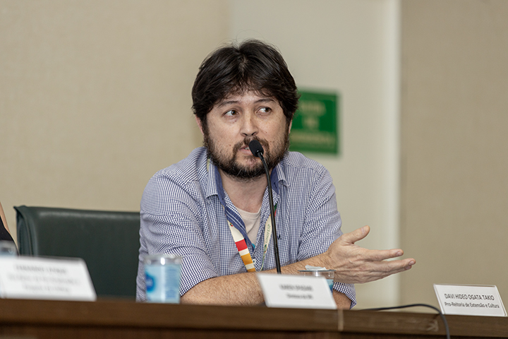 A imagem mostra um homem sentado em uma mesa de conferência ou reunião, falando ao microfone. Ele tem cabelos castanhos escuros, barba, e veste uma camisa xadrez azul-claro com as mangas dobradas. Ele gesticula com a mão direita enquanto fala. Sobre a mesa, há uma placa com seu nome e cargo (aparentemente relacionado a um ministério ou secretaria de cultura), além de uma garrafa d'água e outros materiais. O fundo da sala é neutro, com um tom claro.