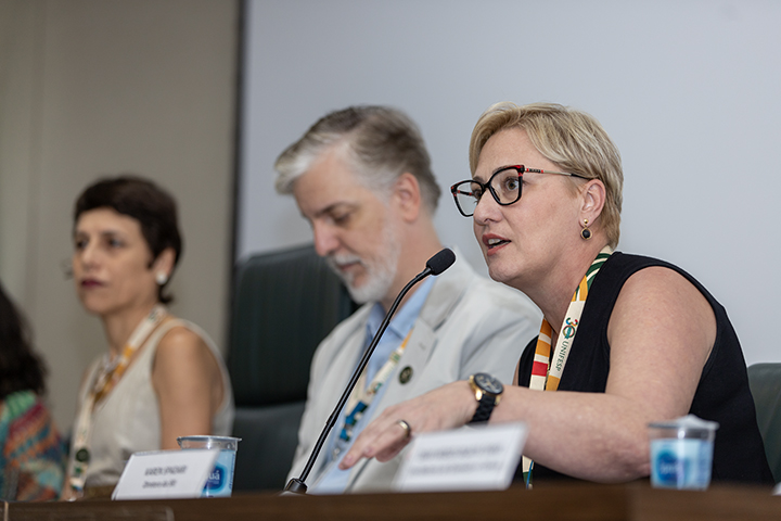 A imagem mostra três pessoas sentadas lado a lado em uma mesa de conferência ou debate. No centro, uma mulher loira de óculos está falando ao microfone. Ela veste roupas formais, usa um colar colorido e parece estar engajada na discussão. À sua direita, há um homem de cabelos grisalhos e barba curta, vestindo um terno claro, que está olhando para baixo. À esquerda, uma mulher de cabelo curto escuro e roupa estampada colorida está observando atentamente. Há plaquinhas com nomes e títulos na mesa à frente de cada pessoa, sugerindo que são palestrantes ou moderadores.