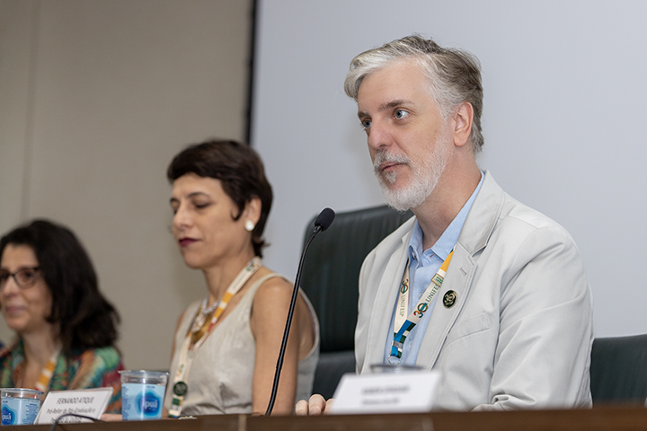 A imagem apresenta três pessoas sentadas em uma mesa de conferência. O foco está em um homem de cabelos grisalhos e barba curta, que veste um terno claro e uma camisa azul. Ele parece atento, olhando ligeiramente para o lado. Ao fundo, à sua esquerda, há duas mulheres: uma com cabelo curto, vestindo uma blusa bege e usando um colar, e outra com cabelo mais longo e óculos, vestindo uma roupa colorida. Elas também estão sentadas à mesa, sugerindo que participam do mesmo evento ou discussão. Há plaquinhas com nomes e copos de água dispostos na mesa, indicando um ambiente formal.