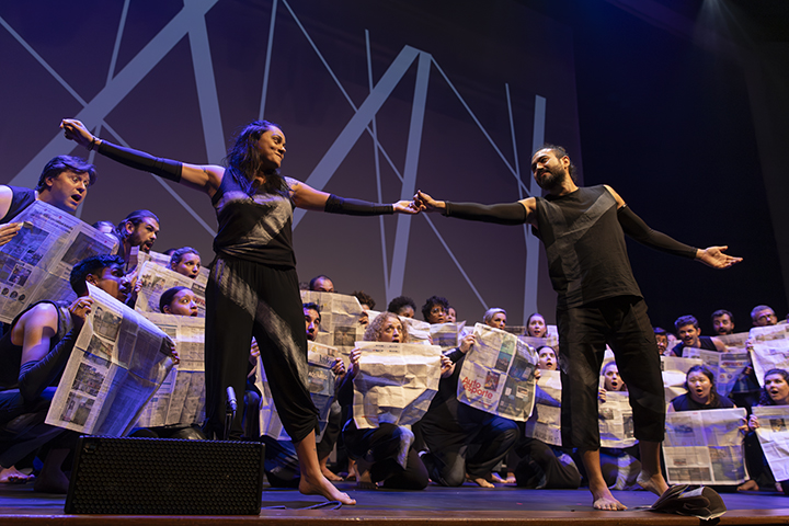 A imagem mostra uma apresentação teatral ou performance no palco. Dois atores estão em destaque na frente, interagindo de mãos dadas. Ambos estão vestidos de preto, com figurinos estilizados. Ao fundo, há um grupo de pessoas segurando jornais abertos, compondo parte da cena. O cenário inclui linhas diagonais no fundo que podem representar uma estética moderna ou abstrata. A iluminação do palco destaca os intérpretes principais e o coro ao fundo.