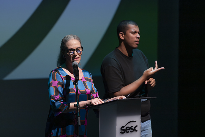 A imagem mostra um evento oficial, possivelmente uma cerimônia ou palestra. À esquerda, uma mulher com óculos e vestido estampado colorido está falando ao microfone, posicionada atrás de um púlpito com o logotipo do SESC. À direita, um intérprete de Libras (Língua Brasileira de Sinais), vestindo camiseta preta e calça jeans, faz gestos para tradução simultânea. O fundo do palco exibe uma projeção em tons de verde e azul com um design abstrato. A iluminação destaca ambos os participantes, criando um ambiente formal e acessível.