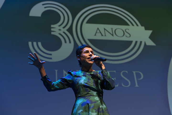 A imagem é um close-up da mesma pessoa no palco, agora destacando seu rosto e parte superior do corpo. Ela continua segurando um microfone com uma das mãos e está gesticulando com a outra, em uma postura expressiva e engajada. No fundo, o logotipo "30 anos" está mais visível, com o texto "ANOS" em destaque dentro de um banner estilizado. A roupa segue sendo um vestido longo com padrões de tonalidades de azul e verde. A iluminação realça tanto a apresentadora quanto o fundo do palco, criando um contraste agradável.