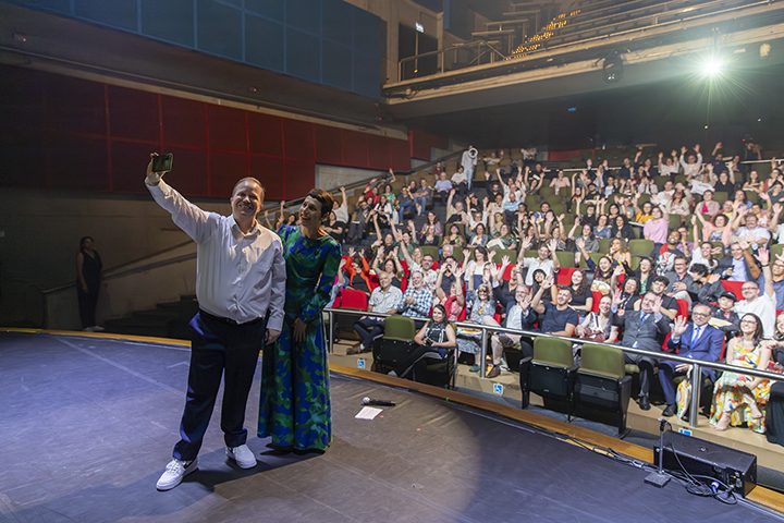 A imagem mostra um momento descontraído durante um evento em um auditório. Duas pessoas estão no palco em destaque: à esquerda, um homem de camisa branca e calça preta tira uma selfie com um celular, enquanto uma mulher à direita, vestindo um vestido longo e estampado em tons de azul e verde, posa sorridente ao seu lado. Ao fundo, a plateia está cheia e aparenta estar animada, com várias pessoas sorrindo, acenando e levantando os braços. A iluminação realça o palco e os dois protagonistas, enquanto o restante do auditório está bem iluminado, destacando a interação calorosa entre os apresentadores e o público.