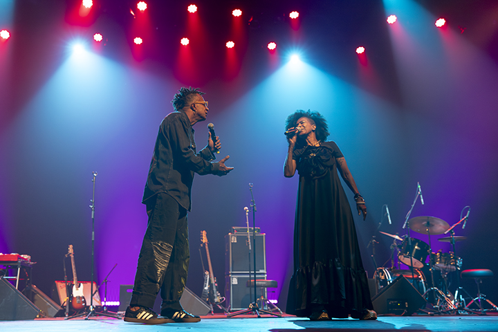 A imagem mostra duas pessoas performando em um palco sob luzes coloridas, com tons de azul, roxo e vermelho no fundo. Ambos estão segurando microfones e parecem estar cantando ou conversando de forma artística.  A pessoa à esquerda veste roupas pretas casuais, incluindo uma camisa e calças com detalhes, e calça tênis preto com listras brancas. Ela faz um gesto expressivo com a mão enquanto interage com a pessoa à direita.  A pessoa à direita usa um vestido longo preto e está de perfil, com uma postura que demonstra envolvimento na performance. Atrás deles, no palco, há instrumentos musicais, como guitarras, bateria e amplificadores, sugerindo um ambiente musical ao vivo. A iluminação cria uma atmosfera vibrante e dinâmica.