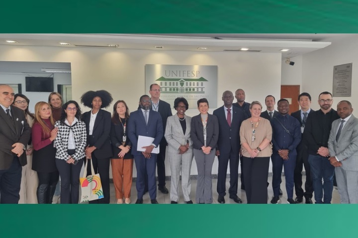 Na imagem, um grupo de pessoas está reunido em frente ao logo da Unifesp, em um espaço institucional, indicando um evento formal. Os participantes estão organizados em duas fileiras e posam para a foto, muitos sorrindo, em um ambiente de celebração ou formalização de um acordo. A diversidade de vestimentas formais e expressões amigáveis demonstra um encontro colaborativo, provavelmente relacionado ao acordo de cooperação técnica entre a Universidade Federal de São Paulo (Unifesp) e o Ministério da Saúde de Angola.  Esse grupo provavelmente inclui representantes da Unifesp e membros da delegação de Angola, como mencionado anteriormente, celebrando a parceria para capacitação e fortalecimento de serviços de saúde em Angola.