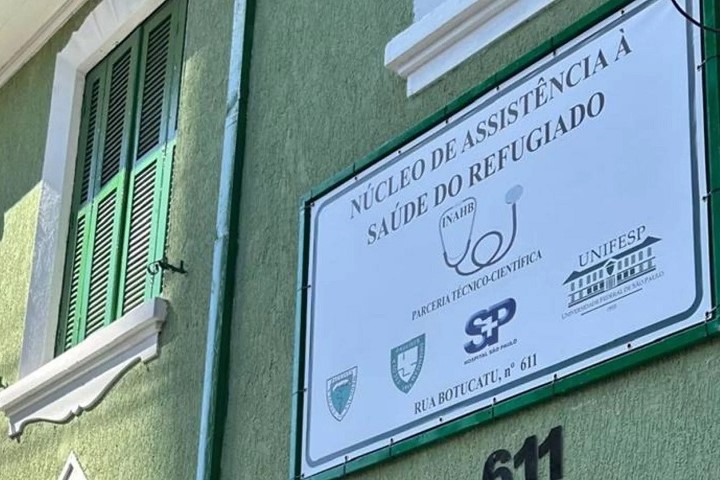 A imagem mostra a fachada de um prédio verde com janelas de madeira pintadas de verde e molduras brancas. Na parede está fixada uma placa branca com bordas verdes, contendo informações sobre uma unidade de atendimento à saúde para refugiados. O texto principal da placa diz: "NÚCLEO DE ASSISTÊNCIA À SAÚDE DO REFUGIADO" Abaixo, há logotipos de instituições envolvidas, incluindo: UNIFESP (Universidade Federal de São Paulo) Governo do Estado de São Paulo Outros logotipos institucionais, incluindo possivelmente faculdades de medicina e entidades de saúde Também está escrito: "PARCERIA TÉCNICO-CIENTÍFICA" "RUA BOTUCATU, nº 611" A placa ainda contém uma imagem de um estetoscópio e a sigla NAHR (provavelmente referindo-se ao núcleo mencionado).