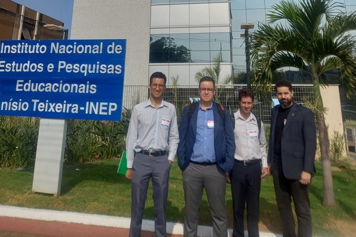 A imagem mostra quatro homens em pé, posando do lado de fora de um prédio, ao lado de uma placa azul que diz: Instituto Nacional de Estudos e Pesquisas Educacionais Anísio Teixeira - INEP. Todos estão vestidos de maneira formal, com camisas sociais e calças, e três deles usam crachás visíveis. Atrás deles, há um prédio com janelas de vidro refletivo e algumas plantas, sugerindo que estão em frente à sede do INEP.  O ambiente parece ser um jardim ou área externa da instituição, com árvores e grama ao redor. A luz do sol sugere que a foto foi tirada durante o dia. O clima é de uma ocasião formal, provavelmente uma visita ou evento relacionado ao INEP.