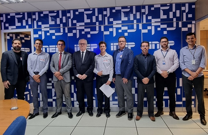 A imagem mostra um grupo de nove homens de pé, posando em frente a uma parede que tem o logotipo do INEP (Instituto Nacional de Estudos e Pesquisas Educacionais). Eles estão organizados em linha, todos vestidos formalmente, com roupas de trabalho, como ternos, camisas sociais e calças. Três deles estão usando crachás visíveis.  Ao fundo, a parede é azul com padrões brancos e as letras INEP destacadas repetidamente. O ambiente parece ser um escritório ou sala de reunião.