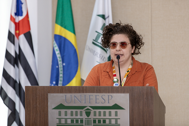 A imagem mostra Bel Moretti Aly, uma pessoa de cabelos curtos e cacheados, usando óculos escuros e camisa laranja. Faz sua apresentação em um púlpito com o logotipo da Unifesp. Ao fundo, estão as bandeiras do Brasil, do estado de São Paulo e da Unifesp.