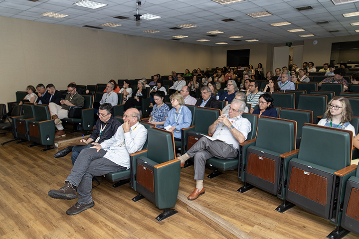 A imagem mostra o público em um auditório durante a audiência pública. As pessoas estão sentadas em fileiras de poltronas verdes, acompanhando atentamente o evento em um ambiente iluminado e organizado.