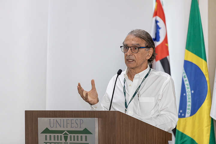 Na imagem, Angelo de Paola, um homem de óculos, cabelos grisalhos e camisa branca. Está fazendo sua apresentação em um púlpito com o logotipo da Unifesp. Atrás dele, há bandeiras do Brasil e do estado de São Paulo.