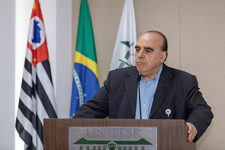 Na imagem Eduardo Medeiros, um homem de terno escuro, camisa azul listrada e cabelos curtos. Faz sua apresentação em um púlpito com o logotipo da Unifesp. Ao fundo, estão as bandeiras do Brasil, do estado de São Paulo e da Unifesp.