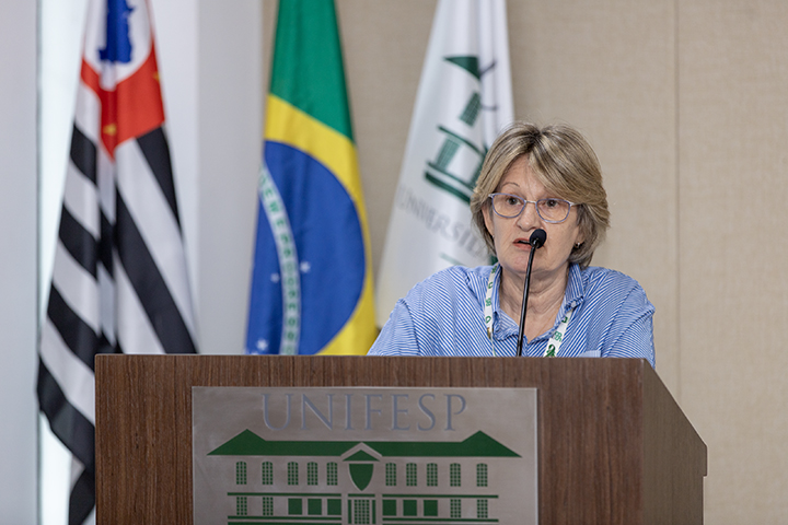 Na imagem Janine Schirmer, uma mulher de óculos, cabelos curtos e loiros, veste uma camisa azul. Faz sua apresentação em um púlpito com o logotipo da Unifesp. Ao fundo, estão as bandeiras do Brasil, do estado de São Paulo e da Unifesp.