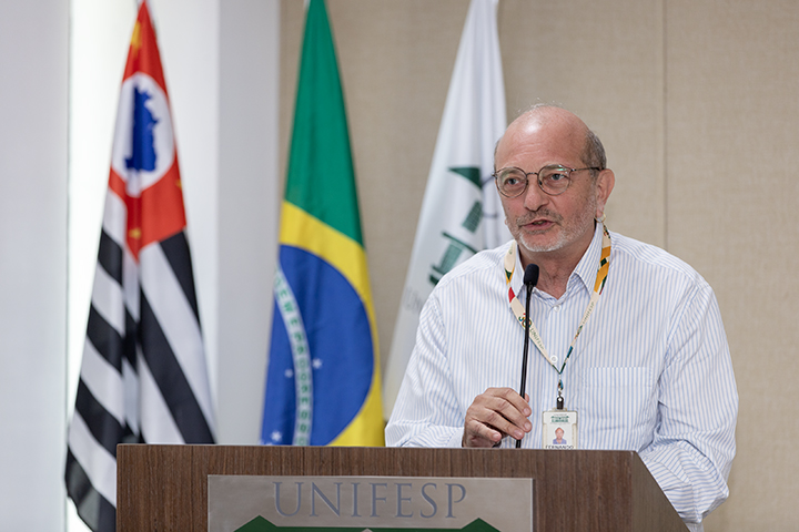 A imagem mostra Fernando Kinker, um homem de óculos, com camisa listrada e crachá. Faz sua apresentação em um púlpito com o logotipo da Unifesp. Ao fundo, estão as bandeiras do Brasil, do estado de São Paulo e da Unifesp.