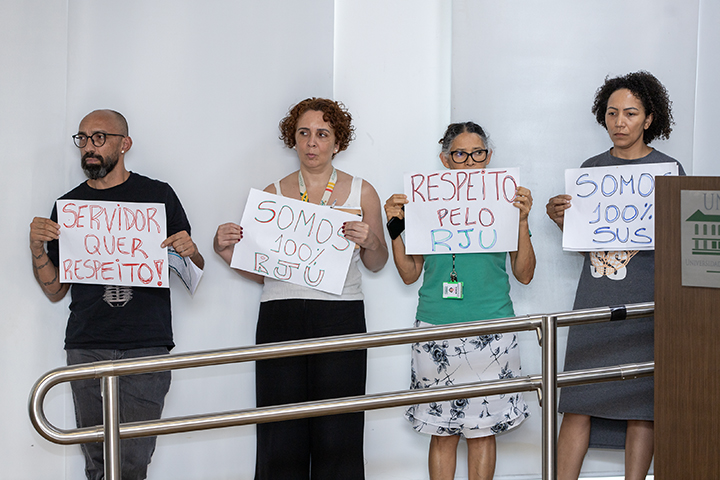 A imagem mostra quatro pessoas segurando cartazes com mensagens como "Servidor quer respeito!", "Somos 100% RJU" e "Respeito pelo RJU". Eles estão em pé ao lado de um corrimão, em um ambiente fechado e iluminado.