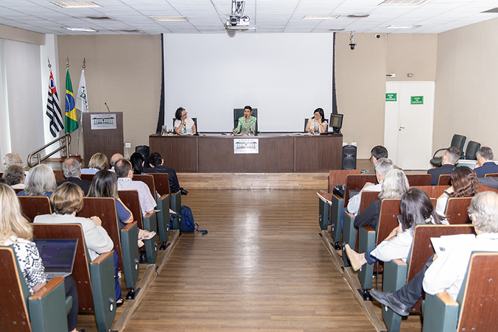 A imagem mostra a audiência pública em um auditório. A vice-reitora, a reitora (que conduz a sessão) e a secretária estão sentadas em uma mesa no palco principal, posicionadas diante de um telão branco. O auditório possui fileiras de cadeiras ocupadas por participantes atentos, incluindo pessoas utilizando laptops. À esquerda, estão bandeiras do Brasil, do estado de São Paulo e da Unifesp. O ambiente é bem iluminado e organizado, com piso de madeira e paredes claras.