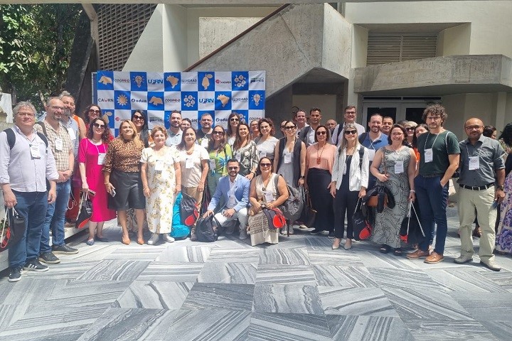 Imagem mostra grupo de pessoas posando para foto em um pátio aberto, na frente de um banner quadriculado em azul e branco