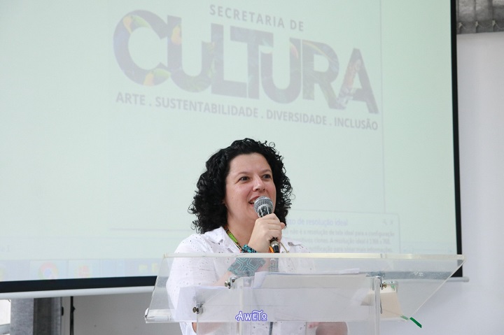 A imagem mostra uma mulher falando ao microfone em um púlpito transparente com o logotipo "Awêlo". Ao fundo, há uma projeção com o texto "SECRETARIA DE CULTURA" e os termos "ARTE · SUSTENTABILIDADE · DIVERSIDADE · INCLUSÃO". A mulher veste roupa branca e acessórios coloridos, sugerindo um discurso institucional em um evento cultural promovido pela Secretaria de Cultura, possivelmente relacionado à valorização dos saberes populares.