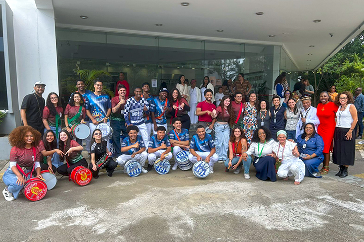 A imagem mostra um grupo diverso de pessoas reunidas em frente a um prédio com uma fachada moderna e envidraçada. Há um destaque para uma bateria de escola de samba, com integrantes vestindo camisetas azuis e brancas e segurando instrumentos de percussão, como surdos, caixas e tamborins. Algumas pessoas estão vestidas de forma casual, enquanto outras apresentam roupas mais formais ou coloridas. O grupo inclui homens, mulheres e jovens de diferentes estilos e idades. O clima parece descontraído e festivo, sugerindo um momento de celebração ou evento cultural. Ao fundo, outras pessoas observam e interagem. A vegetação ao redor complementa o ambiente, que parece ser externo, mas próximo a um espaço institucional.