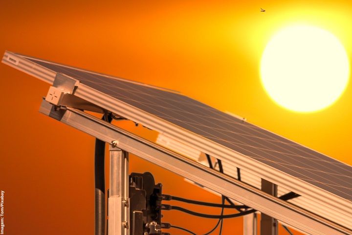  A imagem mostra um painel solar fotovoltaico em destaque contra um fundo alaranjado de um pôr ou nascer do sol. O painel, inclinado para capturar a luz solar, está montado em uma estrutura metálica com fiação visível. A cena evoca um ambiente quente e ensolarado, sugerindo o uso de energia solar como fonte de energia limpa e renovável.