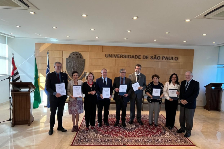  A imagem mostra um grupo de nove pessoas posando para uma foto em um ambiente formal, provavelmente uma cerimônia ou evento institucional. Elas estão em pé, lado a lado, segurando documentos ou certificados nas mãos. Ao fundo, vê-se uma parede com o nome "Universidade de São Paulo" em destaque e o brasão da universidade. À esquerda, estão as bandeiras de São Paulo e do Brasil. O ambiente é bem iluminado, com um tapete vermelho no chão e um púlpito ao lado. As pessoas vestem roupas formais, indicando a importância do evento.