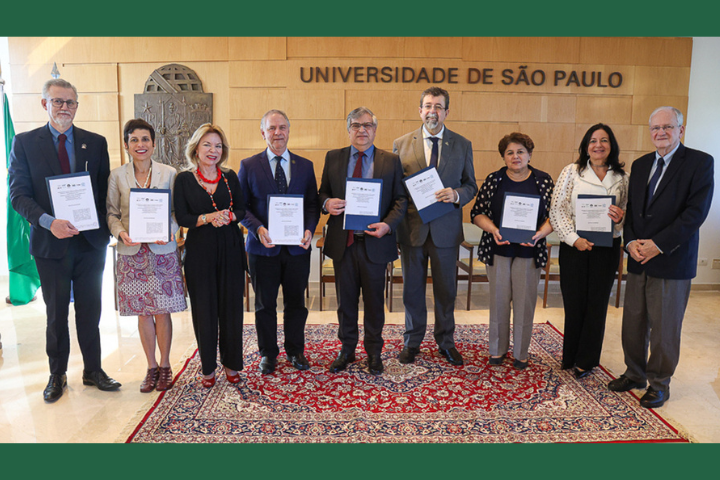  A imagem mostra um grupo de nove pessoas posando para uma foto em um ambiente formal, provavelmente uma cerimônia ou evento institucional. Elas estão em pé, lado a lado, segurando documentos ou certificados nas mãos. Ao fundo, vê-se uma parede com o nome "Universidade de São Paulo" em destaque e o brasão da universidade. À esquerda, estão as bandeiras de São Paulo e do Brasil. O ambiente é bem iluminado, com um tapete vermelho no chão e um púlpito ao lado. As pessoas vestem roupas formais, indicando a importância do evento.