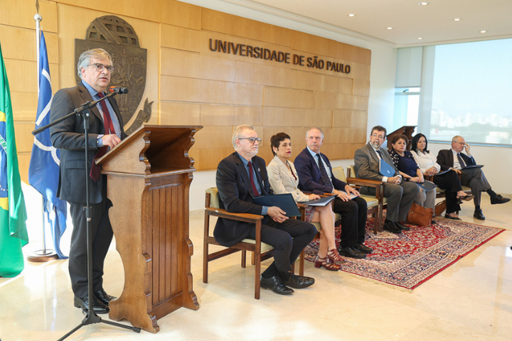 A imagem mostra uma cerimônia formal em um ambiente de reunião na Universidade de São Paulo (USP). No fundo, há um painel de madeira com o brasão e o nome da universidade em letras grandes. À esquerda, uma pessoa está em pé, falando em um púlpito de madeira, enquanto várias outras pessoas estão sentadas em cadeiras ao lado.  Os participantes estão vestidos formalmente, alguns com pastas e documentos nas mãos. Há uma bandeira do Brasil e outra bandeira azul, que pode ser relacionada à universidade, posicionadas ao fundo. No chão, há um tapete persa que complementa a decoração do ambiente, dando um ar solene à cerimônia. A imagem parece registrar um evento oficial ou uma conferência, possivelmente com foco em assuntos acadêmicos ou institucionais.