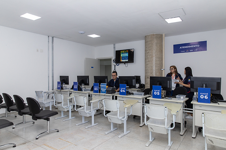 A imagem mostra um ambiente de atendimento ao público, possivelmente em uma farmácia ou setor de saúde. O espaço é composto por um balcão de atendimento com seis guichês numerados de 01 a 06, cada um equipado com monitores de computador e cadeiras para funcionários e clientes. Há funcionários uniformizados no ambiente: um deles está sentado no Guichê 04, enquanto outros dois estão em pé, conversando ou organizando algo próximo ao Guichê 06.  Na parede ao fundo, há uma placa azul com a palavra 
