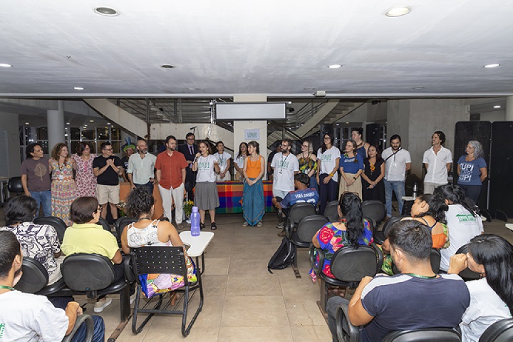A imagem mostra um evento ou apresentação em um espaço interno, provavelmente em uma universidade ou centro cultural. Há um grupo de aproximadamente 20 pessoas de pé na frente, enfileiradas, voltadas para o público. Essas pessoas aparentam ser organizadores ou palestrantes. Algumas delas usam camisetas com logotipos ou nomes de instituições, como "UNB" e "UPI", sugerindo uma relação com universidades. No público, diversas pessoas estão sentadas em cadeiras giratórias, observando atentamente a apresentação. As roupas coloridas de algumas pessoas da plateia indicam diversidade cultural, possivelmente com participação de comunidades indígenas ou tradicionais. Há também uma mesa ao fundo coberta com uma toalha colorida, e um banner com o logotipo "LINDI" ao centro