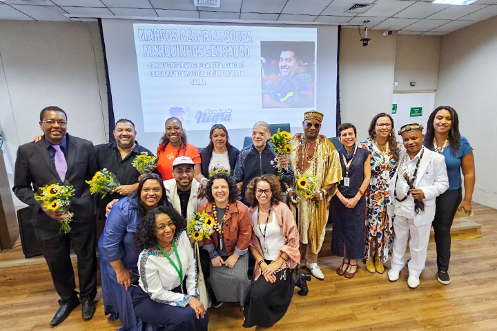 A imagem mostra um grupo de pessoas posando para uma foto em um auditório. O grupo inclui homens e mulheres de diferentes idades e estilos, e alguns seguram buquês de girassóis, sugerindo um momento de celebração ou homenagem. Ao fundo, há uma projeção com texto que inclui o nome "Marcos César de Sousa - Marquinhos Sensação", possivelmente referindo-se a um homenageado ou tema do evento. Também há logotipos e menções a uma "Comunidade Negra".  Entre os participantes, há diversidade de roupas, incluindo trajes formais e tradicionais, como uma vestimenta dourada com influência africana e colares de contas. A cena reflete um evento cultural ou acadêmico, destacando a celebração de identidades e realizações afrodescendentes. A atmosfera é de alegria e reconhecimento.