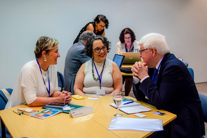 A imagem mostra um grupo de pessoas em um ambiente de reunião formal, provavelmente uma conferência ou encontro institucional. Em destaque, três pessoas estão sentadas à mesa conversando de forma animada e cordial. São duas mulheres e um homem (de cabelos brancos), todos com crachás pendurados em cordões azuis no pescoço. Ao fundo, outras pessoas também trabalham.