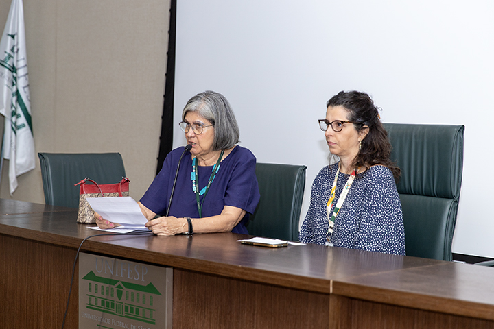 A imagem mostra duas mulheres sentadas em uma mesa de conferência ou painel. A mulher à esquerda está lendo um documento em voz alta, enquanto a outra, à direita, observa atentamente. Ambas estão usando crachás, sugerindo que participam de um evento ou reunião oficial. O cenário parece ser formal, possivelmente em uma instituição, pois há uma bandeira e um logotipo na mesa que parece indicar uma universidade ou órgão acadêmico. A mulher à esquerda usa óculos, tem cabelos grisalhos e veste uma blusa azul. A mulher à direita tem cabelo castanho preso, também usa óculos e veste uma blusa com estampas geométricas. A mesa à frente delas tem o logotipo da "Universidade Federal de Santa Catarina" (UFSC).