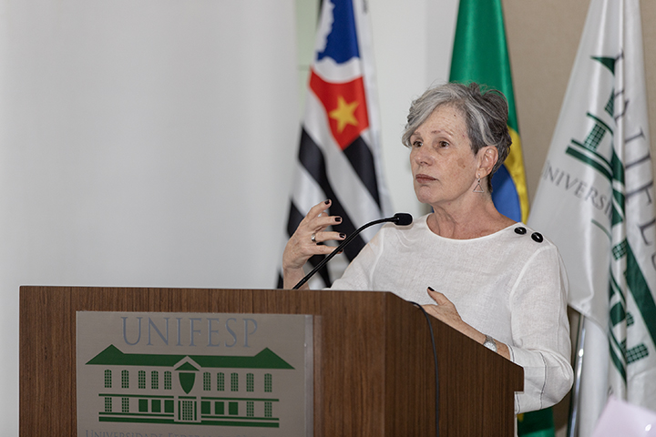 A imagem mostra uma mulher falando em um púlpito, possivelmente durante uma apresentação ou palestra. Ela tem cabelos grisalhos curtos e está usando uma blusa clara. Enquanto fala, faz gestos com as mãos para enfatizar seu ponto. Atrás dela, há várias bandeiras, incluindo a bandeira do Brasil e outra que parece ser do estado de São Paulo. O púlpito tem o logotipo da "UNIFESP" (Universidade Federal de São Paulo), o que indica que o evento ocorre nessa instituição. A expressão da palestrante é séria e focada, sugerindo que ela está discutindo um tópico importante.