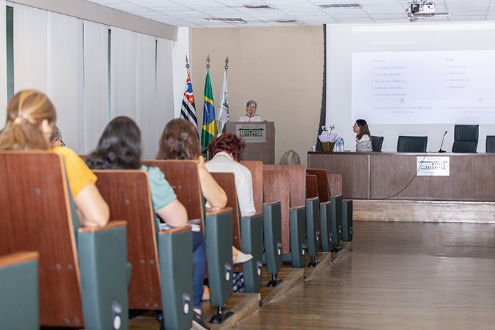 A imagem mostra uma sala de conferências com várias pessoas sentadas em cadeiras voltadas para a frente, onde uma mulher está em um púlpito fazendo uma apresentação. Ao fundo, há uma projeção na parede com texto, sugerindo que a palestrante está discutindo tópicos específicos ou exibindo uma apresentação de slides. A palestrante está posicionada em frente a três bandeiras: a do Brasil, a do estado de São Paulo e outra possivelmente da instituição. O púlpito traz o logotipo da UNIFESP (Universidade Federal de São Paulo). À direita da imagem, uma outra pessoa está sentada à mesa, observando a apresentação. A sala tem um ambiente formal, com cadeiras estofadas em tom de marrom e um piso de madeira. As pessoas na plateia parecem atentas ao que está sendo dito.