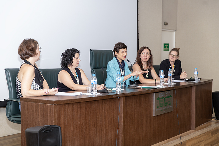 A imagem mostra uma mesa de conferência ou seminário com cinco mulheres participando. Elas estão sentadas atrás de uma mesa de madeira com microfones, copos de água e papéis. O logotipo da UNIFESP (Universidade Federal de São Paulo) está visível na parte frontal da mesa. O ambiente parece ser um auditório ou sala de eventos acadêmicos.