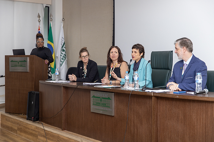 A imagem mostra uma mesa de debate ou conferência em um auditório, com cinco pessoas visíveis. Quatro delas estão sentadas atrás de uma mesa equipada com microfones, garrafas de água e documentos. Uma pessoa adicional está de pé em um púlpito à esquerda, utilizando um notebook.Detalhes específicos:Participantes sentados na mesa:Da esquerda para a direita, há três mulheres e um homem.Uma delas está falando ao microfone, gesticulando.Os participantes estão formalmente vestidos, sugerindo um evento oficial ou acadêmico.Bandeiras ao fundo:Há duas bandeiras visíveis, sendo uma do Brasil e outra de uma instituição.Pódio à esquerda:A pessoa no púlpito parece estar monitorando ou controlando a apresentação.Logotipo e nome da instituição:A mesa e o púlpito têm o logotipo da UNIFESP (Universidade Federal de São Paulo), indicando que o evento ocorre nesta instituição.O cenário sugere um evento formal, como uma conferência acadêmica, uma mesa-redonda ou um debate.