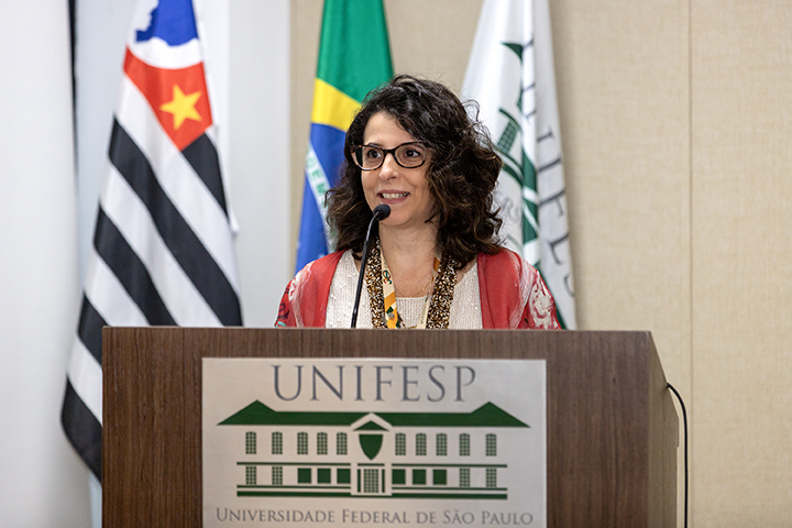 A imagem mostra Ana Gouw, a pró-reitora de graduação da Unifesp, falando em um púlpito. Ela está sorrindo e usando óculos, com cabelos castanhos escuros e ondulados. Veste um casaco vermelho sobre uma blusa clara. Ao fundo, há bandeiras do Brasil, do estado de São Paulo e da própria Unifesp. No púlpito, o logo da Universidade Federal de São Paulo (Unifesp) está visível. Ana parece estar discursando em um evento oficial da universidade, com um microfone à sua frente.