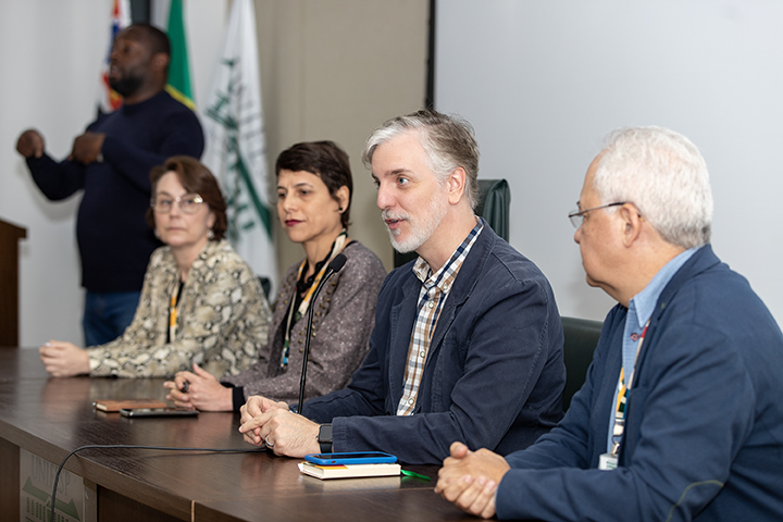 Na imagem, os quatro membros da mesa de abertura, acompanhadas por um intérprete de Libras ao fundo. Eles parecem concentrados na fala de Fernando Atique , com bandeiras ao fundo, incluindo a do Brasil.