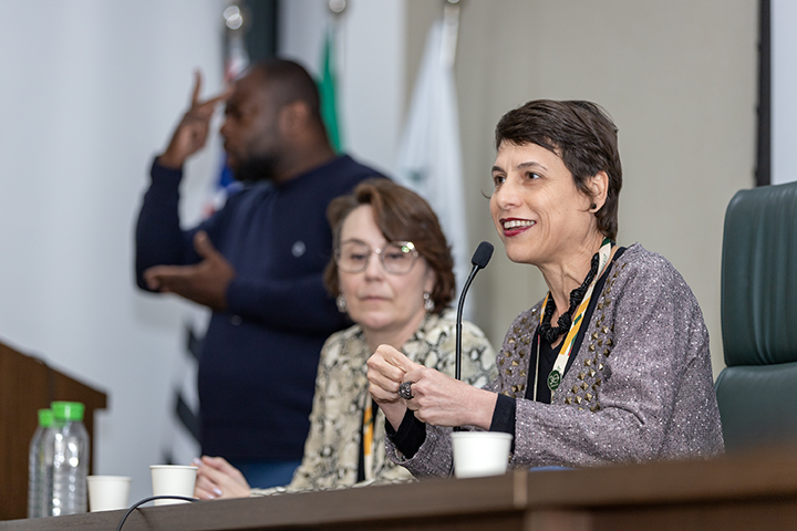 A foto mostra duas mulheres sentadas em uma mesa. À direita, Raiane Assumpção está falando ao microfone com um sorriso, gesticulando levemente com as mãos. Ela usa uma blusa de manga comprida em tons claros e um crachá. À sua esquerda, Gabriela Arantes Wagner está sentada, observando com uma expressão atenta. Ela usa óculos e veste uma blusa estampada. Na mesa à frente delas, há garrafas de água e copos descartáveis. Ao fundo, um intérprete de Libras está em pé, gesticulando, e algumas bandeiras estão parcialmente visíveis. O ambiente parece ser formal e voltado para uma apresentação.