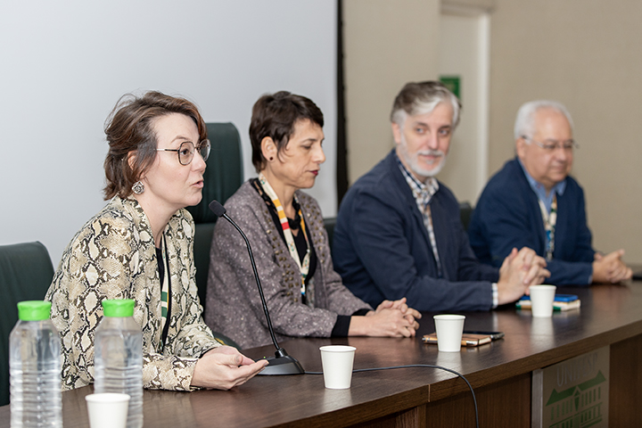 Na imagem, Gabriela Arantes Wagner fala ao microfone, enquanto Raiane Assumpção, Fernando Atique e Elbert Macau, sentados ao seu lado, a observam atentamente. Todos estão em uma mesa de madeira, onde há copos descartáveis e garrafas de água. O ambiente é formal, sugerindo um evento ou conferência, com os participantes concentrados na discussão.