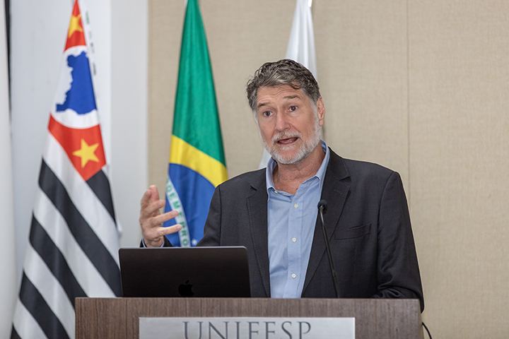 A imagem mostra Jean Pierre Henry Balbaud Ometto falando em um púlpito, identificado pela inscrição "Unifesp". Ele tem cabelos grisalhos e barba curta também grisalha. Está usando um terno escuro e uma camisa social azul clara. Em segundo plano, há duas bandeiras: a da bandeira do estado de São Paulo à esquerda e a do Brasil ao centro. O homem parece estar gesticulando com a mão direita enquanto fala. Está dando uma palestra.