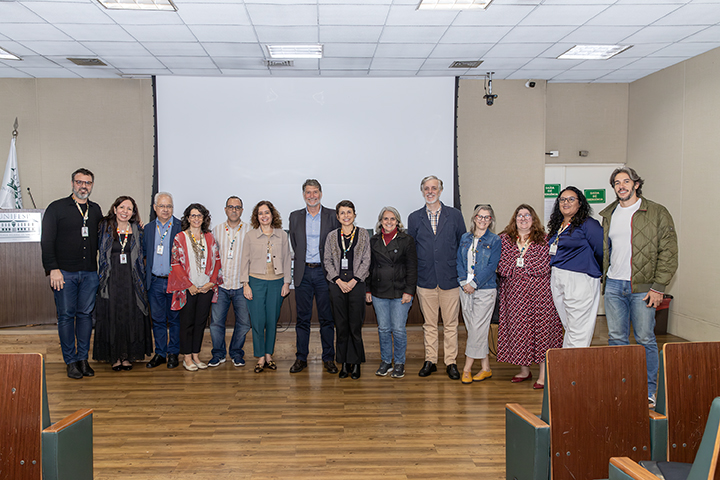 A imagem mostra um grupo de membros da Unifesp em pé, lado a lado, em uma sala de conferências com piso de madeira e cadeiras em estilo de auditório. Ao fundo, há uma tela de projeção branca e o teto é composto de placas brancas com luminárias. Todas as pessoas na imagem parecem estar participando de um evento formal, algumas vestindo crachás. A maioria está vestida de forma casual, com jaquetas, camisas e calças. Eles posam para a foto de maneira organizada, formando uma linha horizontal, aparentando estarem envolvidos com um evento acadêmico ou profissional.