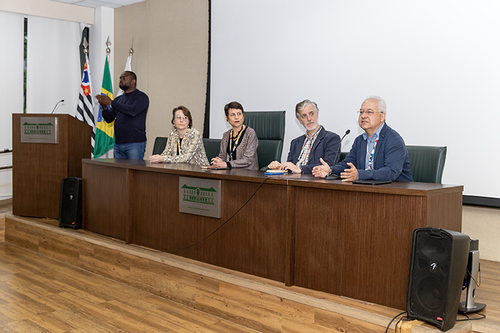 A imagem mostra uma mesa de abertura com quatro pessoas sentadas em um auditório. Da esquerda para a direita: Gabriela Arantes Wagner, Raiane Assumpção, Fernando Atique e Elbert Macau. Todos estão sentados atrás de uma mesa de madeira escura com o logotipo da Unifesp. Eles usam crachás e parecem participar de uma discussão formal ou apresentação. Ao lado esquerdo da mesa, um homem em pé, à frente de um púlpito, um intérprete de Libras. No fundo, há bandeiras do Brasil, do estado de São Paulo e da Unifesp. A sala tem piso de madeira, e uma grande tela de projeção branca está atrás dos participantes.