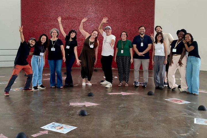 A imagem mostra um grupo de 12 pessoas posando juntas de forma descontraída em frente a uma parede vermelha curva, em um espaço amplo e iluminado. Algumas pessoas fazem gestos animados com as mãos, como acenos ou sinais de vitória, enquanto outras sorriem diretamente para a câmera, criando um clima de descontração e celebração.  No chão, há objetos hemisféricos pretos e folhas com imagens ou desenhos coloridos espalhados, sugerindo que o local é parte de uma instalação artística ou atividade coletiva. O grupo parece estar em um momento de confraternização ou encerramento de uma experiência em grupo, todos usando crachás que indicam participação em um evento organizado. A disposição das pessoas e o ambiente sugerem uma interação criativa e colaborativa.