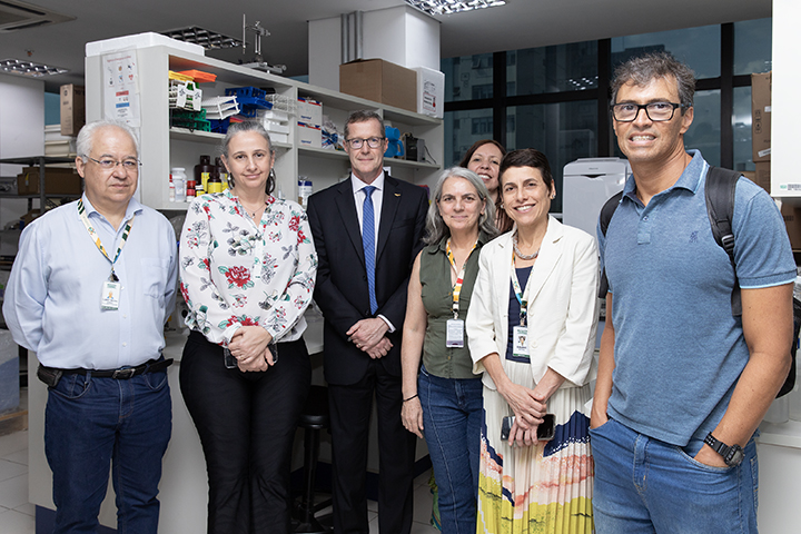 A foto mostra sete pessoas em pé, lado a lado, em um ambiente de laboratório com prateleiras ao fundo. Todos estão sorrindo, alguns usam crachás e jalecos, em uma visita as instalações do Campus São Paulo.