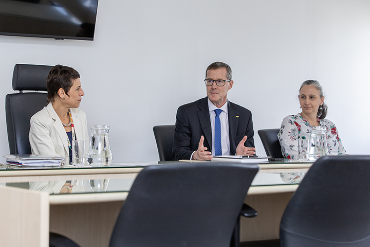 A imagem mostra a reitora da Unifesp, o reitor e a vice-reitora do ITA  em uma reunião formal, sentadas à mesa em um ambiente iluminado. O homem ao centro gesticula enquanto fala, enquanto as mulheres ao seu lado escutam atentamente.