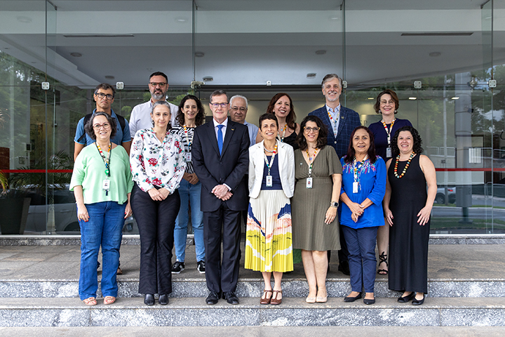 A foto mostra um grupo de 15 pessoas posando juntas em frente a um prédio com portas de vidro. O grupo está organizado em duas fileiras: uma na frente, em pé sobre degraus, e outra atrás, na mesma altura do chão. Todos estão vestidos formalmente ou casualmente elegantes, e muitas das pessoas usam crachás pendurados no pescoço. O ambiente parece ser uma entrada corporativa ou institucional, com pisos de granito e um local bem iluminado. Ao fundo, há portas de vidro e uma área interna parcialmente visível.
