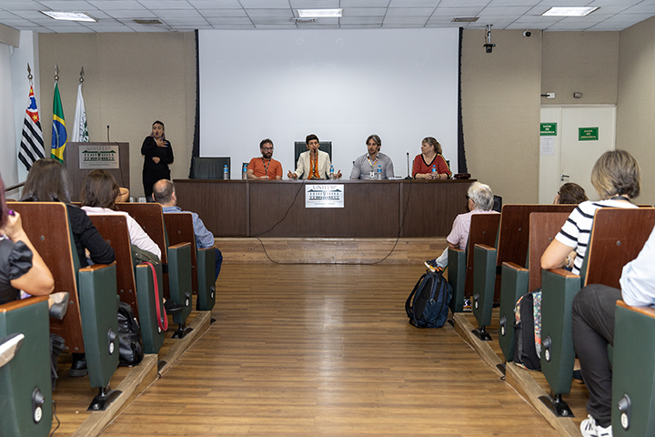 Imagem mostra anfiteatro térreo da Reitoria da Unifesp, com pessoas sentadas na plateia e as pessoas que compõem a mesa de abertura do evento ao centro