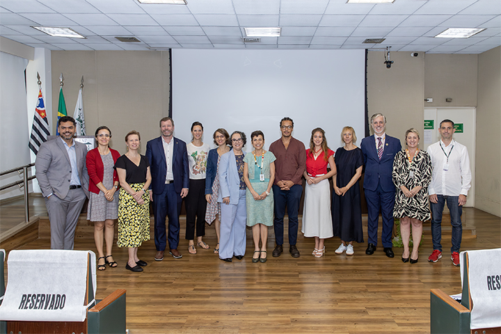 A imagem mostra um grupo de 13 pessoas em pé, organizadas em uma fileira no que parece ser um auditório ou sala de conferências. O grupo é composto por homens e mulheres de diferentes idades, vestindo roupas formais e semiformais. Ao fundo, há uma tela de projeção grande, ainda sem exibição de conteúdo. Também podem ser vistas algumas bandeiras à esquerda da imagem, entre elas a bandeira do estado de São Paulo e a bandeira do Brasil, sugerindo um contexto oficial ou institucional.